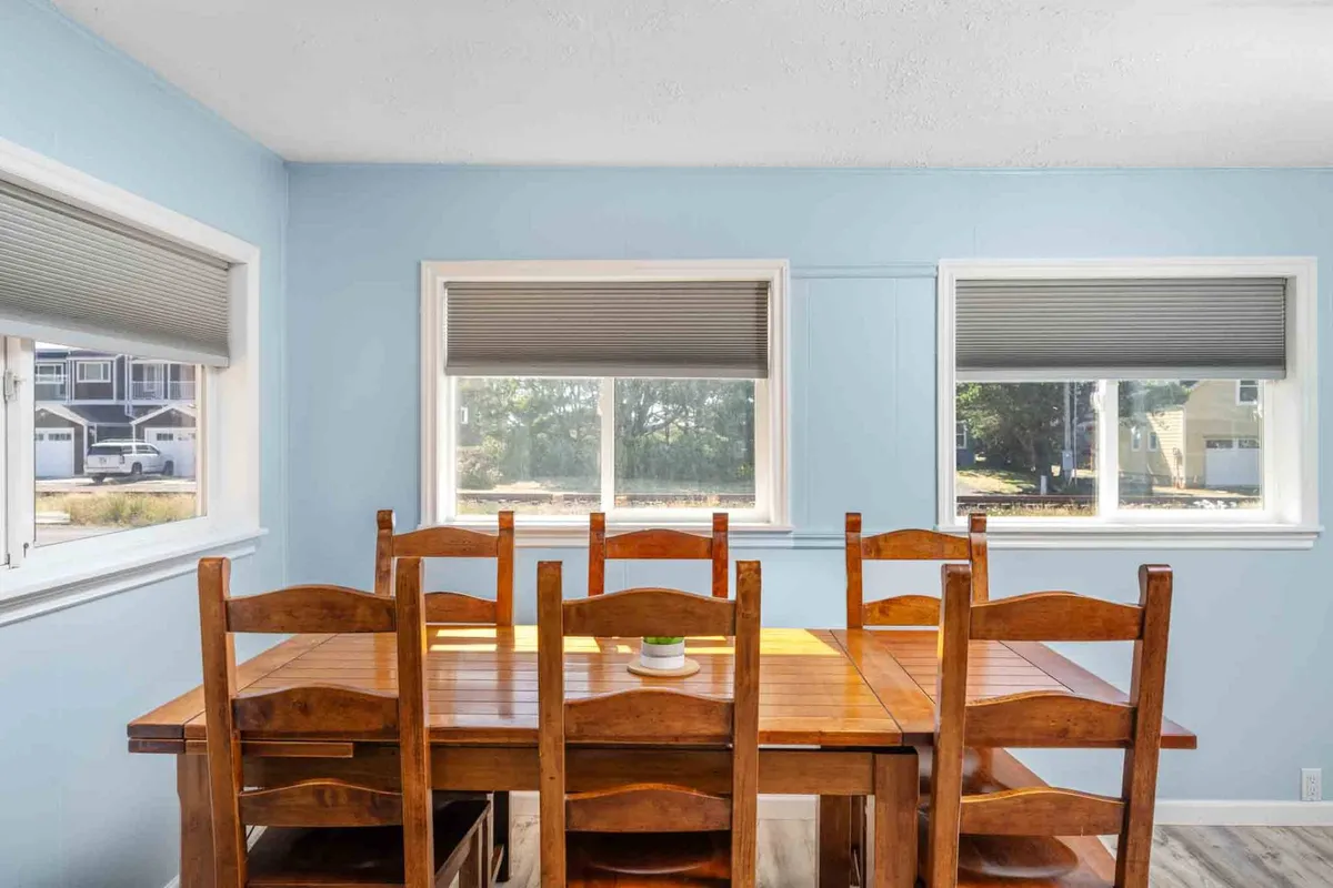 A spacious dining area featuring a wooden dining table surrounded by comfortable chairs and bathed in natural light from large windows. The setting is perfect for family-style meals, social gatherings, and lively conversation.