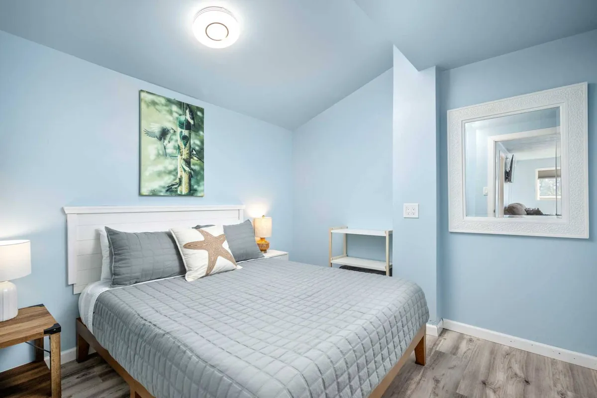 A serene bedroom with a neatly made bed, soft lighting from a bedside lamp, a decorative mirror, and wall art that adds personality. The space exudes tranquility and comfort, inviting guests to relax, read, or unwind.