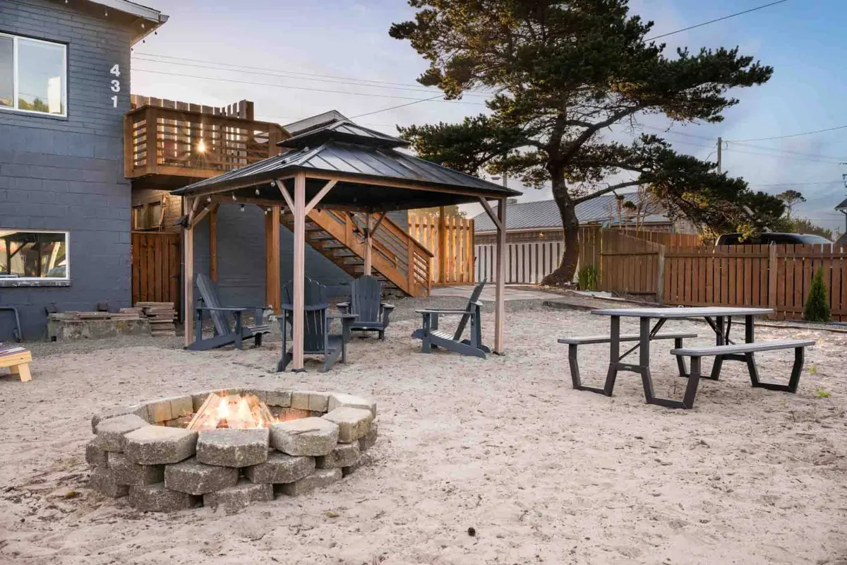 An inviting exterior space with a prominent fire pit, outdoor seating area, wooden deck, and a gazebo in the background. The ambiance is warm and communal, ideal for evening gatherings, enjoying meals outdoors, or relaxing by the fire as the sun sets.
