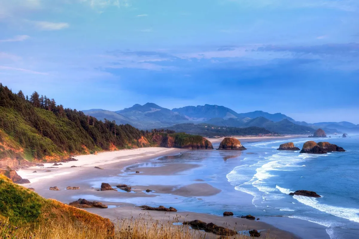 A breathtaking view of the ocean with waves rolling onto a sandy beach, set against a backdrop of rock formations, forests, and distant mountains. The tranquil scene invites exploration and highlights the proximity to coastal attractions.