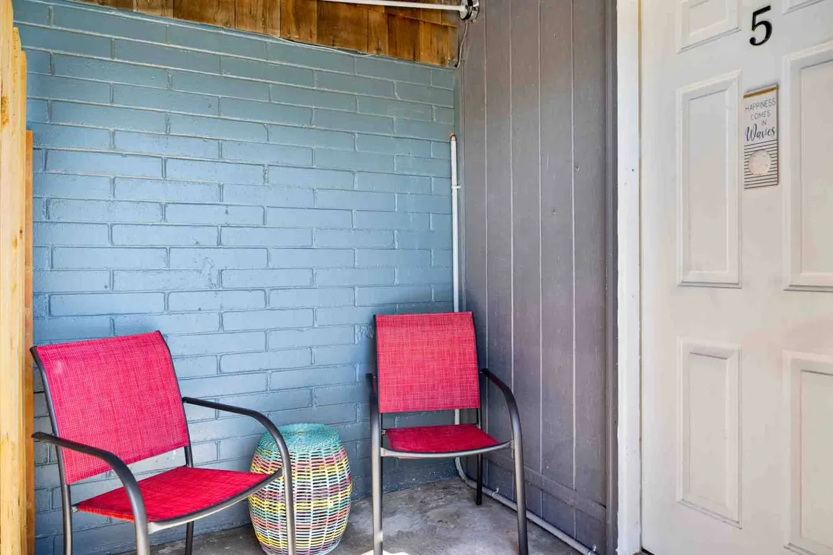 An inviting patio area featuring red chairs and a colorful stool, positioned outside a door labeled with the number 5. The scene is bathed in natural sunlight, creating a tranquil spot perfect for savoring a morning coffee or enjoying an intimate conversation. The relaxed ambiance highlights the privacy and peacefulness of this outdoor nook.