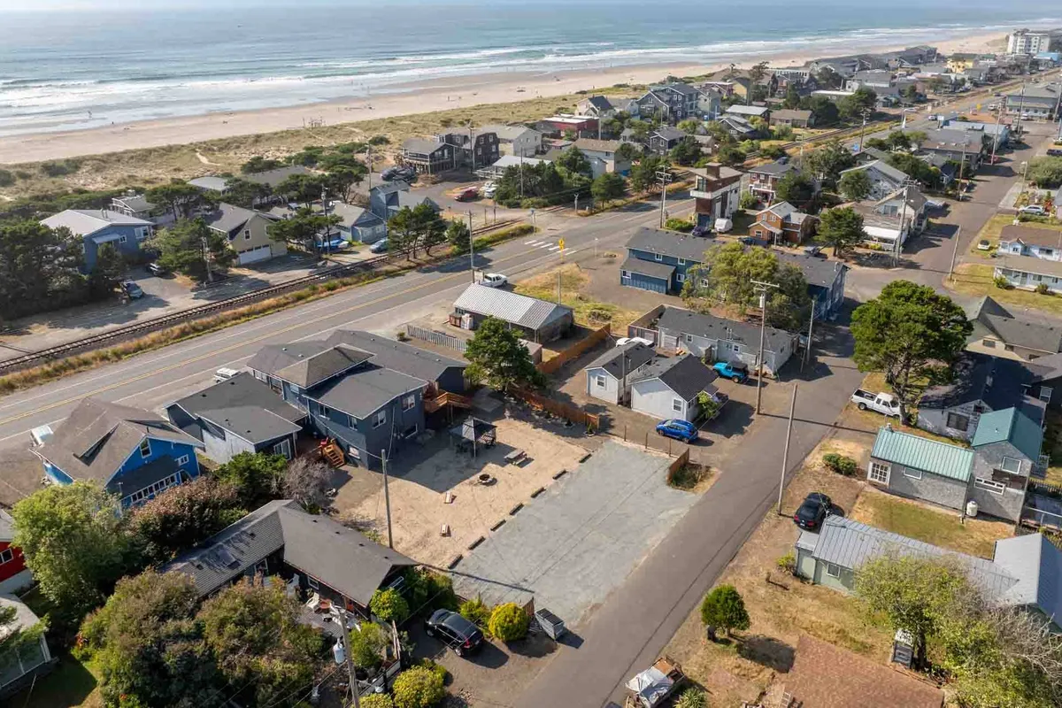A relaxed beachfront scene showing sandy shoreline, neighborhood houses just steps away, a tree-lined roadway, and plenty of open sky. The setting suggests easy access for walks along the beach and exploring the local area, perfect for guests with pets.