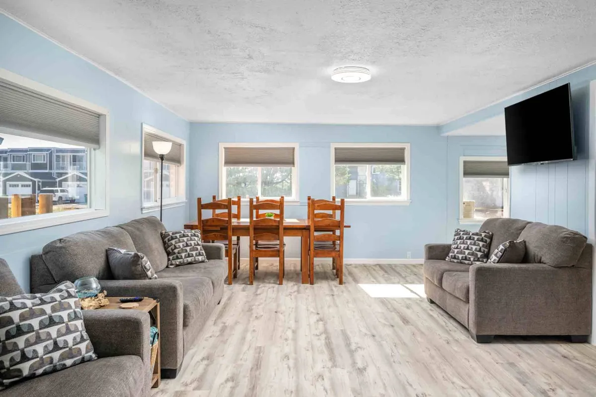 A cozy open-plan living and dining space featuring plush sofas, a central dining table, large windows allowing natural light to fill the room, and a television. The arrangement encourages both relaxation and social gatherings, and the open layout highlights modern, comfortable furnishings.