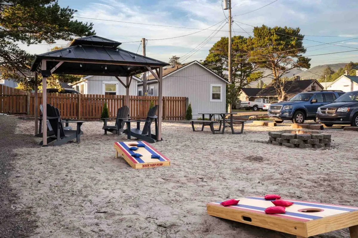 A spacious communal outdoor area featuring a gazebo, cornhole game, fire pit, and ample seating. The setup encourages social interaction, relaxation, and play, with the gazebo acting as a hub for gatherings or enjoying the sunset.