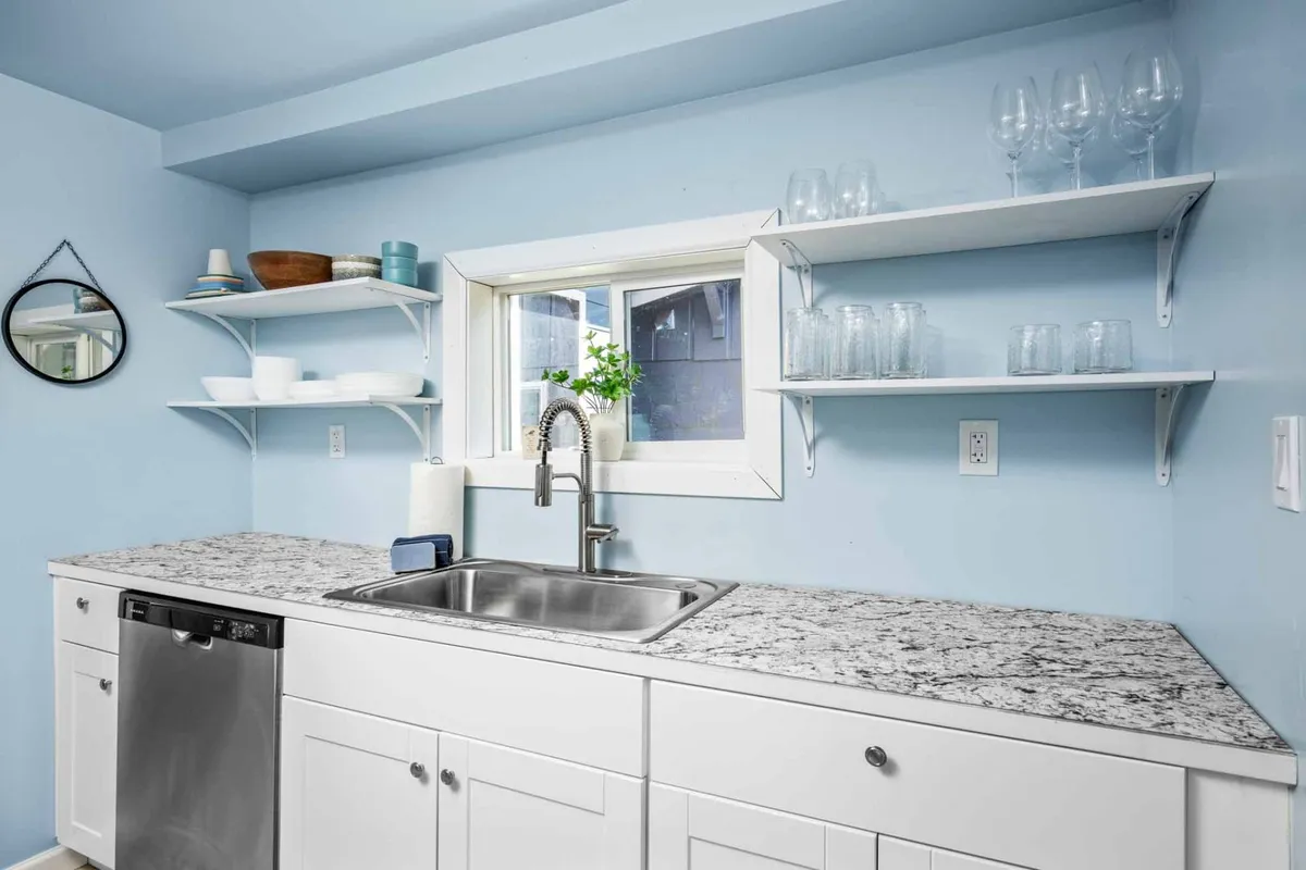 A cheerful kitchen nook with a granite countertop, sink, shelves stocked with dishes, and a window adorned with potted plants. Natural light streams in, creating a cozy space for meal prep and casual conversation.