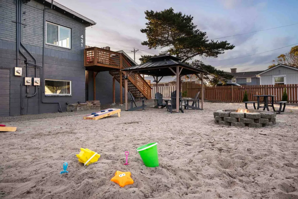 A family-friendly exterior scene showing a sand play area with toys, a gazebo, fire pit, and comfortable lawn furniture. Children are playing in the sand while families gather nearby, suggesting an atmosphere of relaxation and togetherness after a day at the beach.