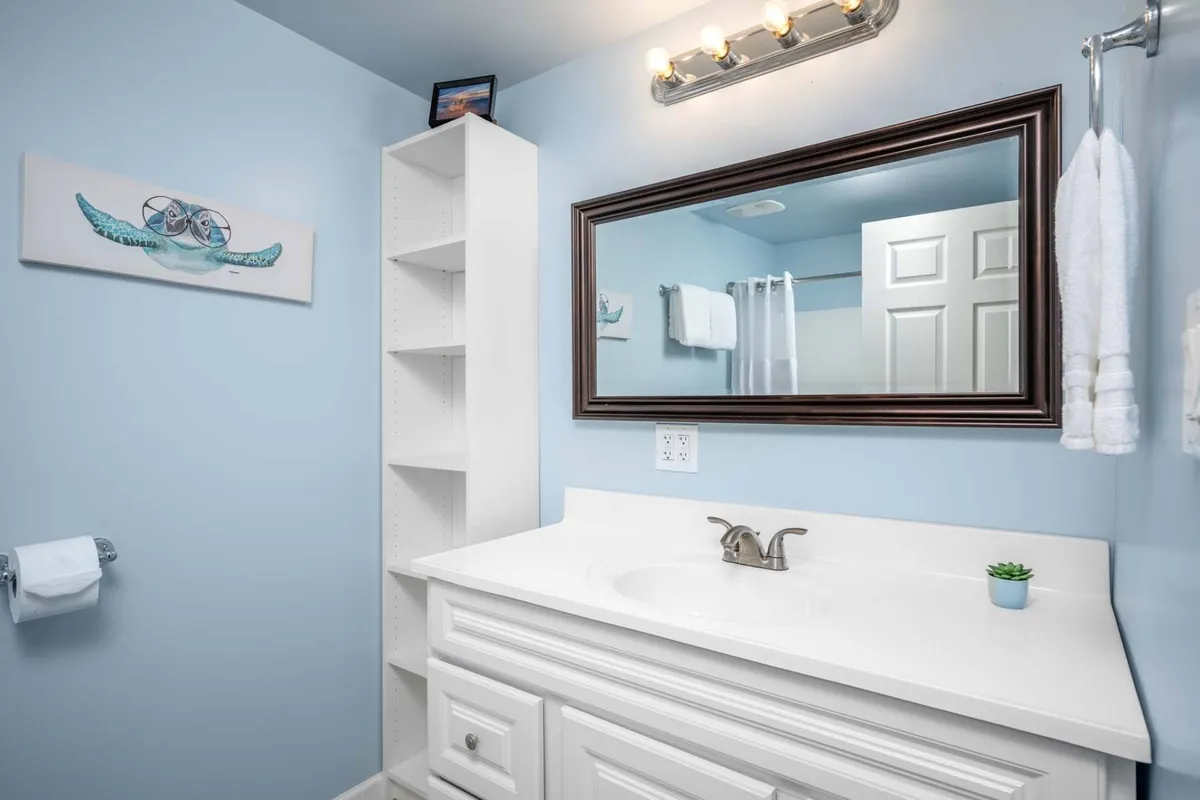 A modern bathroom featuring a crisp white vanity, a large mirror, blue accent walls, a shelf unit, and thoughtful decorative details. The space is bright, inviting, and designed for self-care routines.